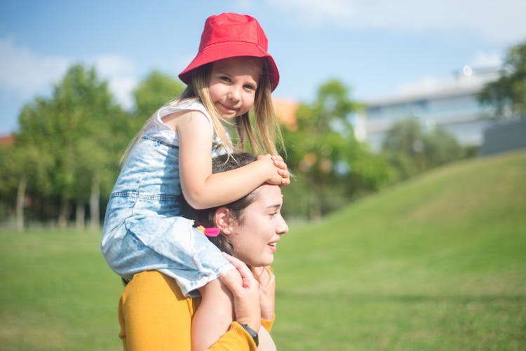 Woman In Yellow Long Sleeve Shirt Carrying Girl With Red Bucket Hat On Shoulders