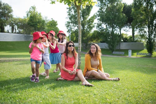 Women Sitting on Green Grass Field Beside Girls Playing