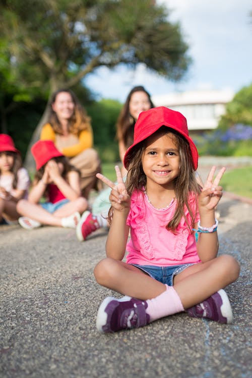 Girl in Pink Shirt with Red Bucket Hat Sitting on Gray Concrete Pavement Making a Peace Sign