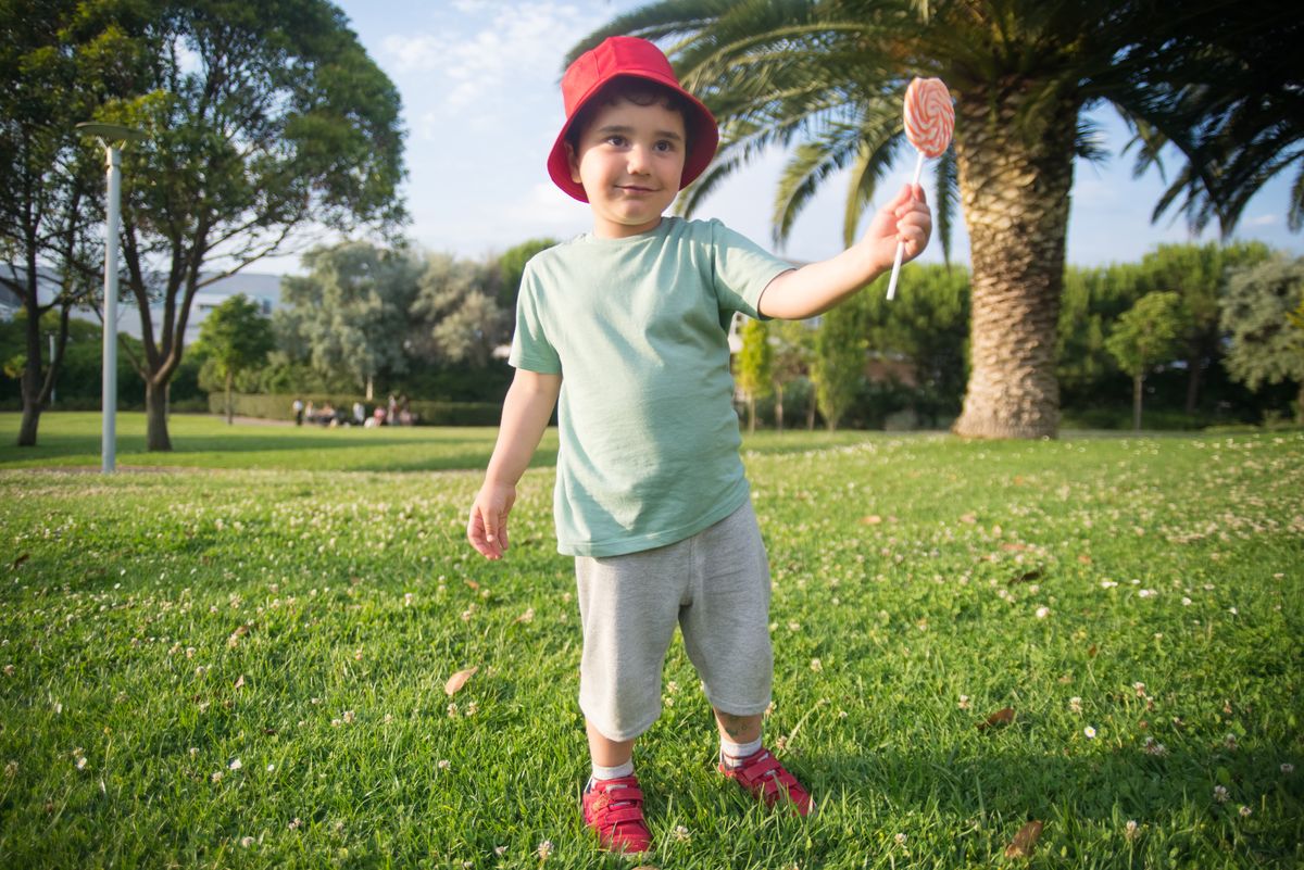 Happy child in a park holding a lollipop, wearing a red hat and smiling on a sunny day.
