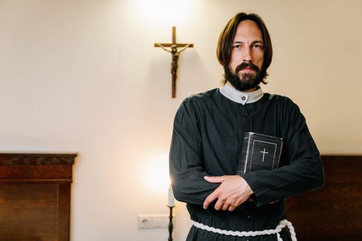Bearded clergyman in religious attire holding a Bible indoors near a cross.