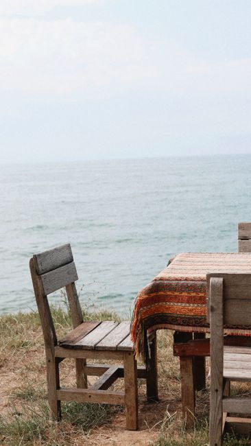 Rustic wooden chairs and a table with a patterned cloth by the ocean, perfect for relaxation or gatherings.