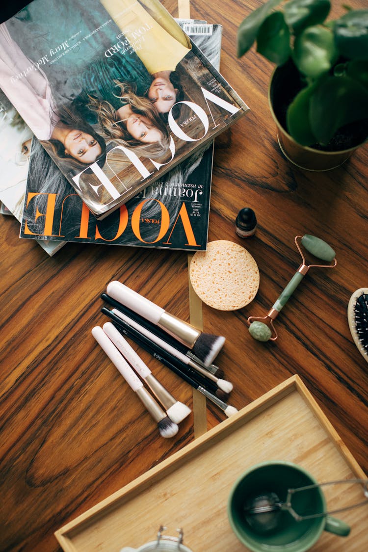 Vogue Magazines On Wooden Table With Makeup Brushes And Jade Roller