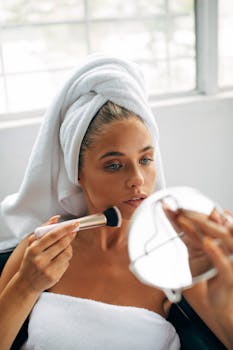 Caucasian woman applying makeup with brush after bath, wrapped in towel.