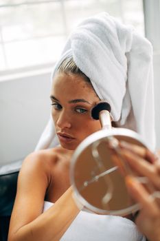 Caucasian woman in towel applying makeup with a brush, holding mirror.