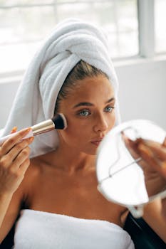 Caucasian woman in towel applying makeup with brush in front of mirror.
