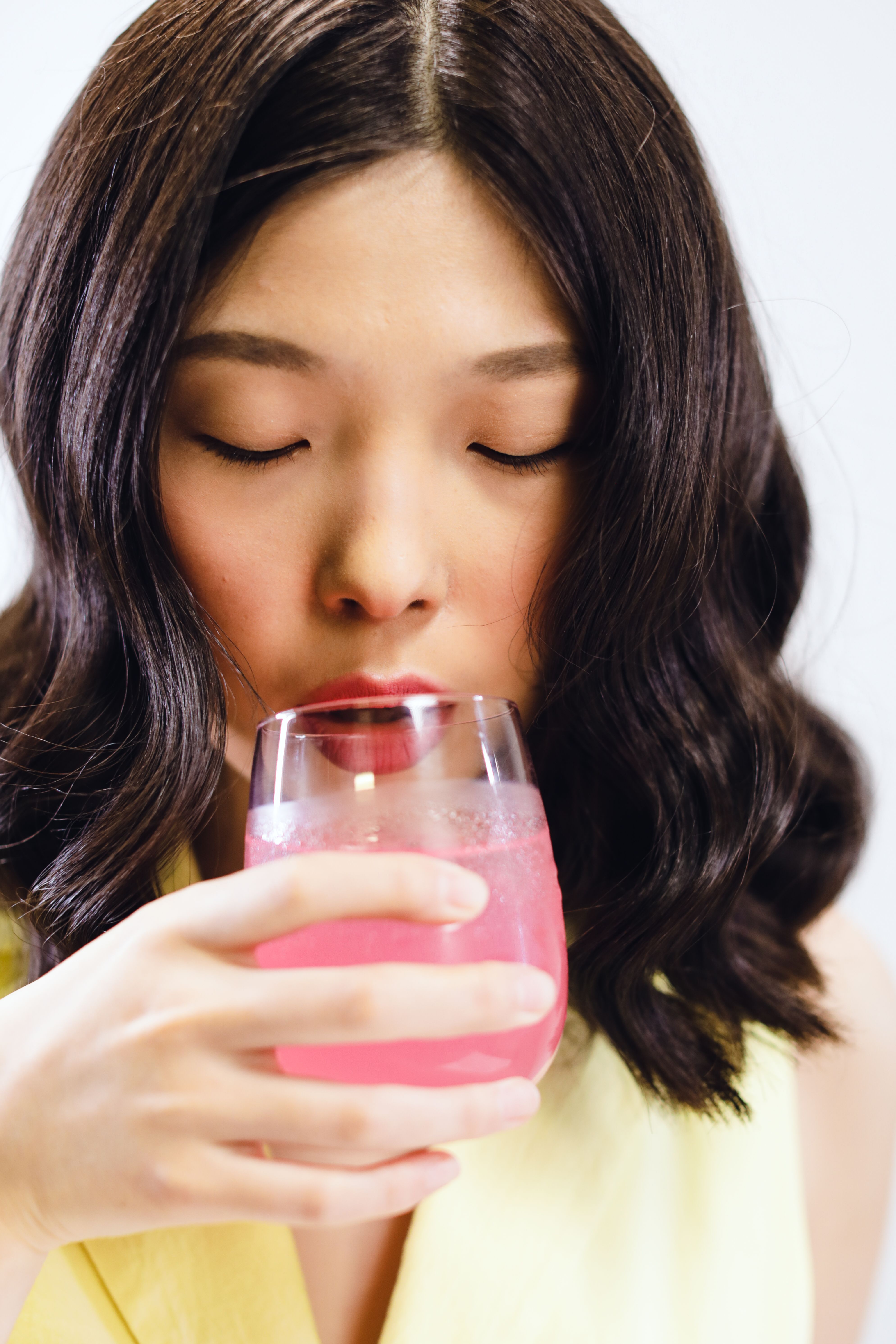 Asian woman savoring a vibrant pink beverage indoors with eyes closed, capturing a moment of refreshment.