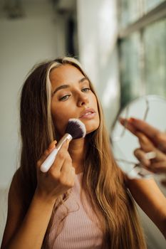 Young woman using a makeup brush in front of a mirror indoors.
