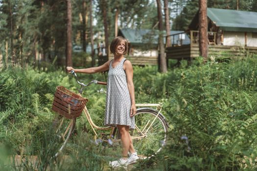 Smiling woman in summer dress poses with bicycle in lush forest setting.