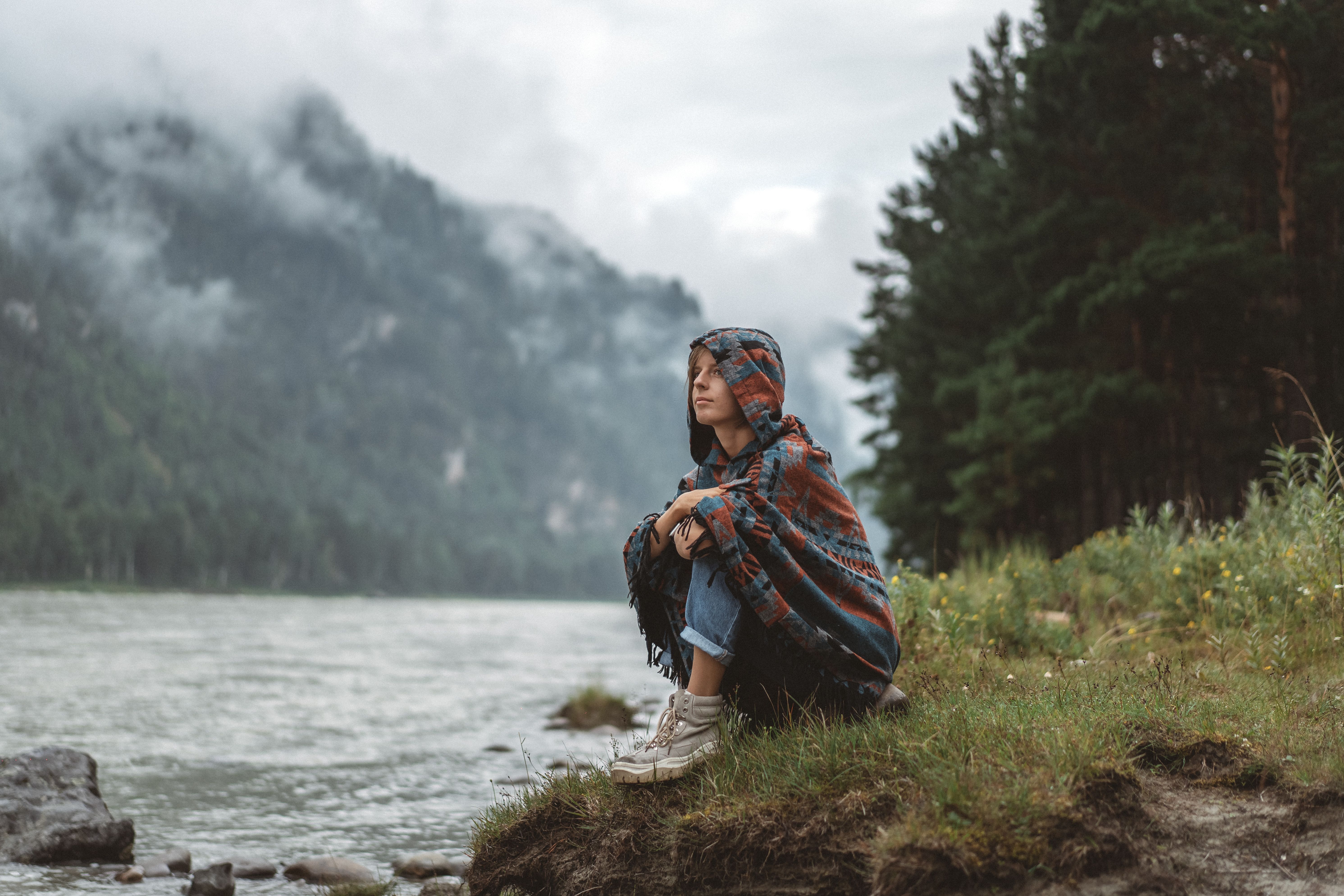 Woman in a patterned blanket sits by a misty river, embracing tranquility.