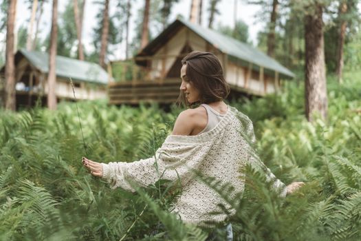 A woman in a white cardigan explores a tranquil forest with wooden cabins, enjoying the serene greenery.