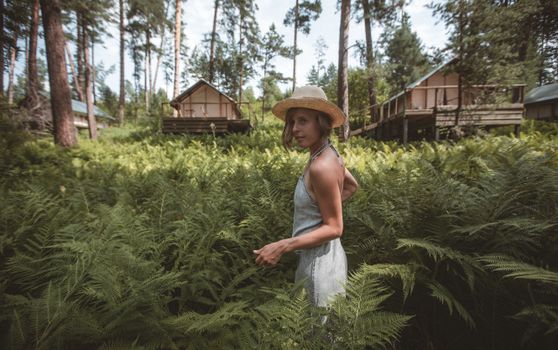 Woman wearing a straw hat stands among fern plants in a forest with cabins in the background.