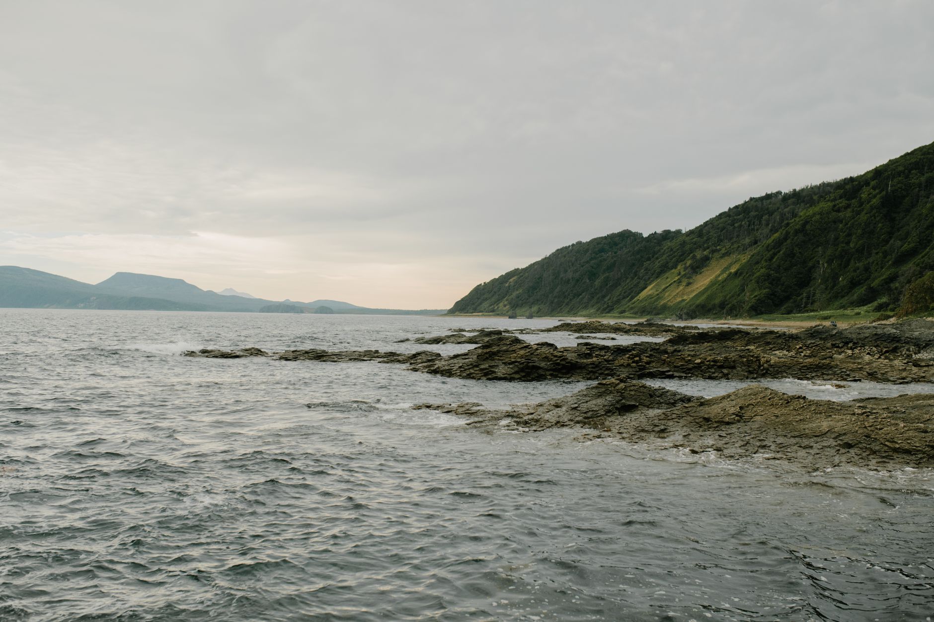 A serene view of the Sakhalin coastline with rocky shores and lush hills under an overcast sky.