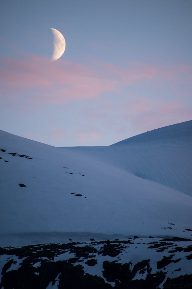 A Half Moon On Blue Sky Over A Snow Capped Mountain