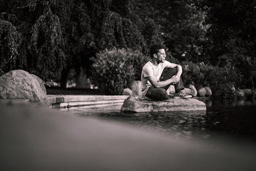 Free Grayscale Photo of Man Sitting on Rock Surrounded by Water  Stock Photo
