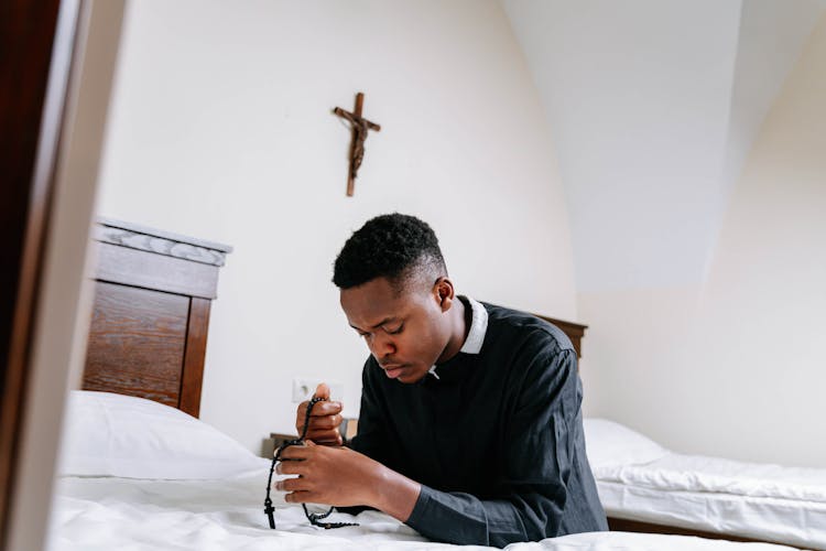 Man Kneeling In Prayer Holding A Rosary Inside His Bedroom