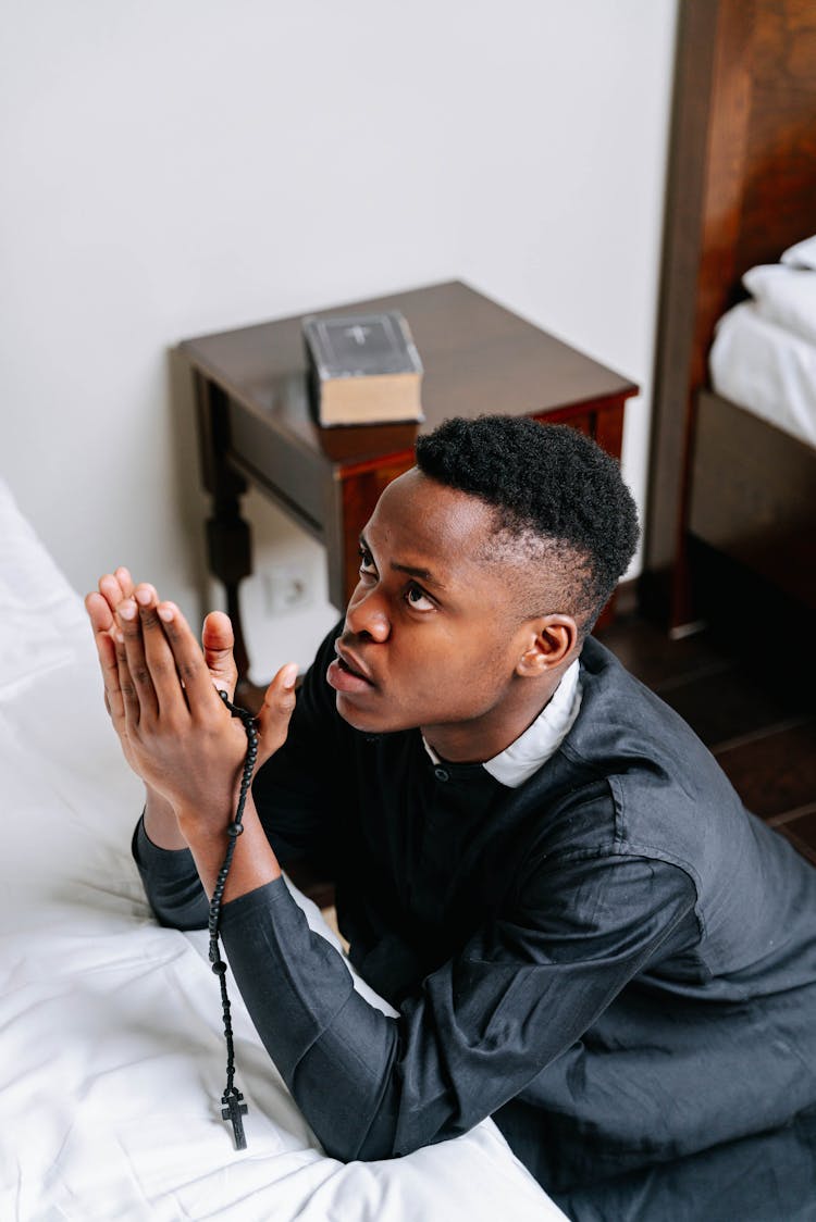 Man Kneeling In Prayer Holding A Rosary Inside His Bedroom