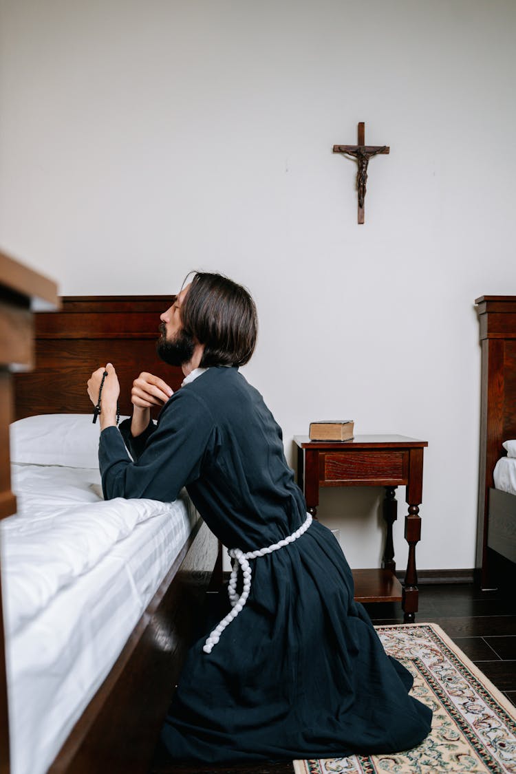 Man Holding A Rosary And Kneeling In Prayer Inside A Bedroom