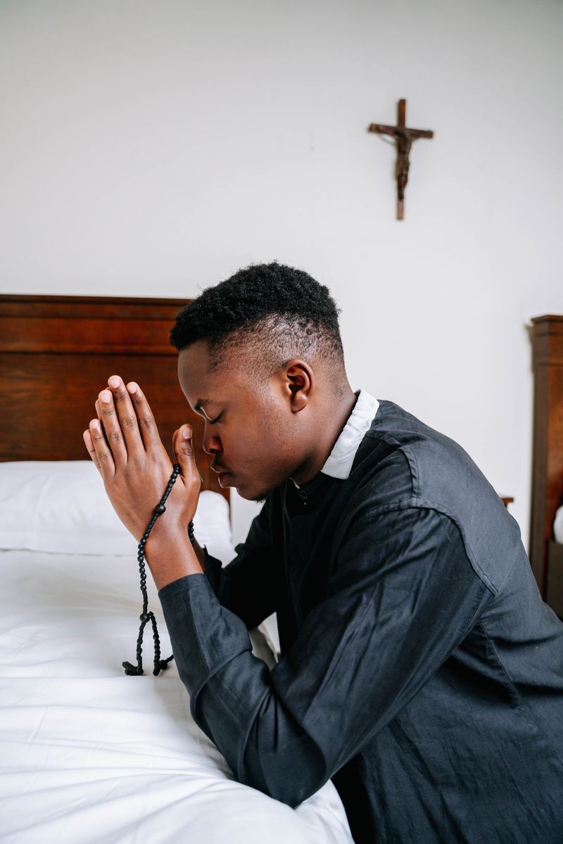 A young man kneels by his bed praying with a rosary in a peaceful bedroom setting, emphasizing faith and reflection.