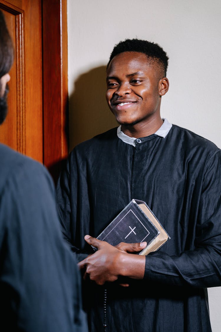 Man In Black Clergy Robe Holding A Holy Bible And Prayer Beads Having Conversation With His Friend