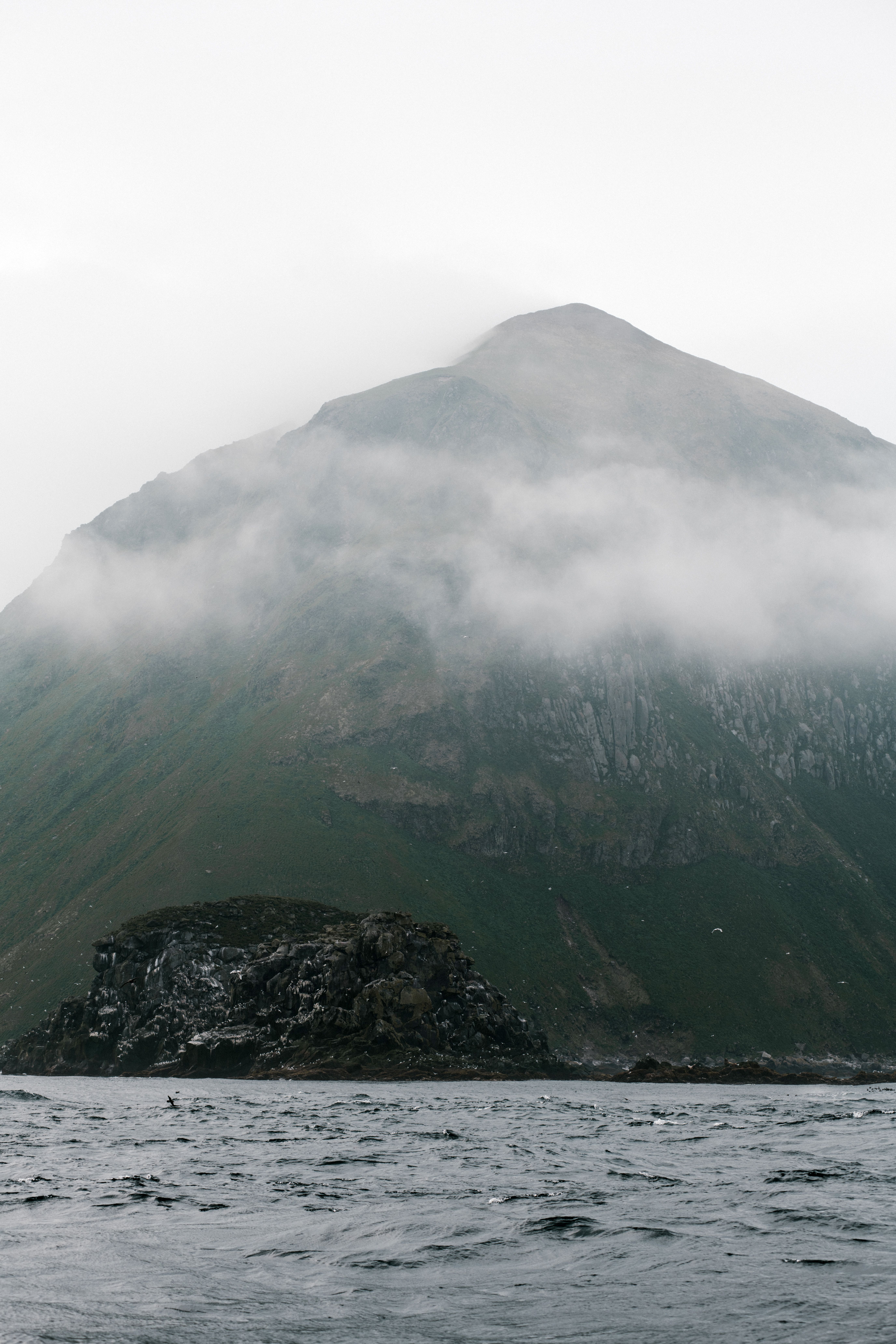 Dramatic view of a misty mountain partially covered by fog with a rocky sea shore.