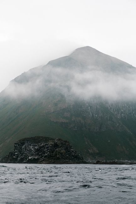 Dramatic view of a misty mountain partially covered by fog with a rocky sea shore.