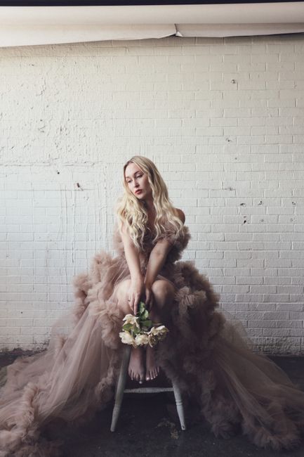 A beautiful woman in a tulle gown sits barefoot holding a bouquet in front of a white brick wall.