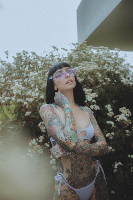 Tattooed woman in a bikini posing outdoors with flowering plants, wearing sunglasses.