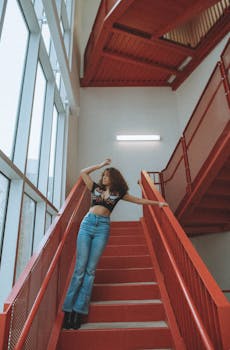 A fashionable adult woman stands elegantly on a red staircase indoors, showcasing modern style and architecture.