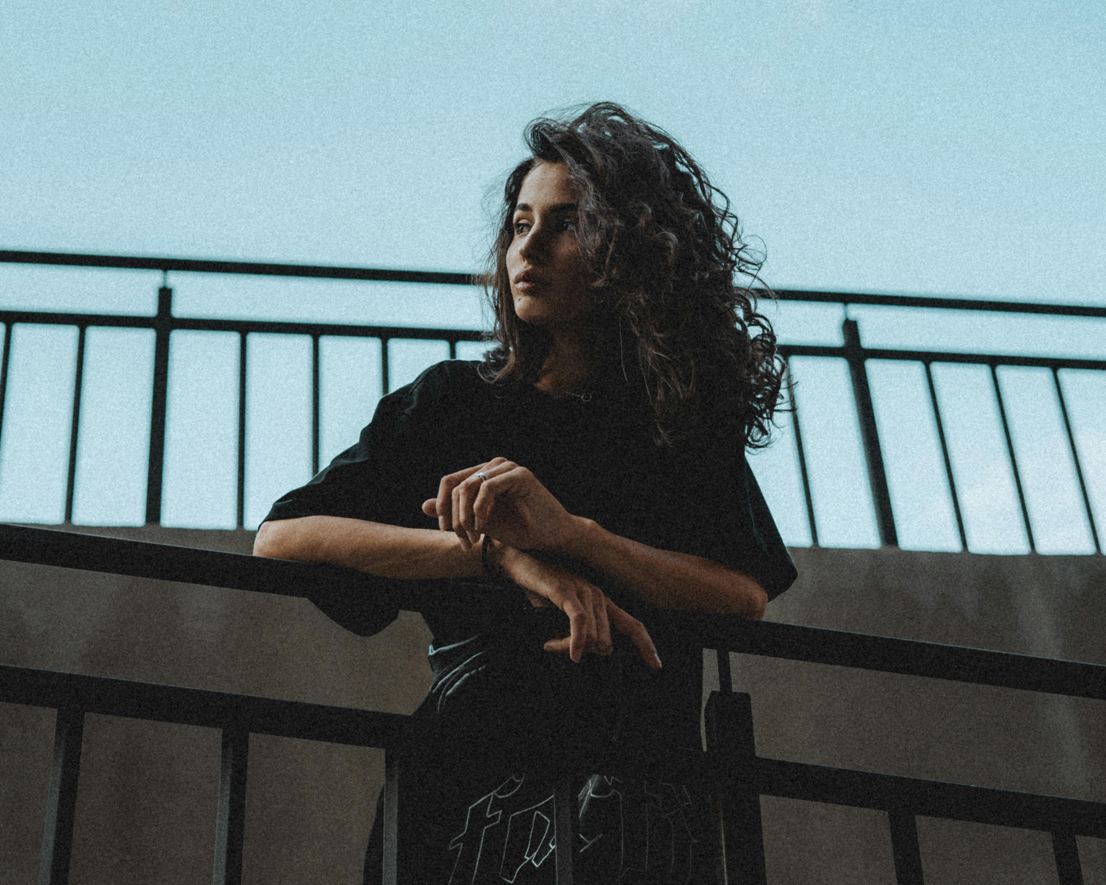 A fashionable woman with curly hair stands pensively against a railing under the clear sky.