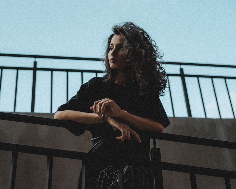 Low angle of attractive young female with curly hair in stylish clothes leaning on hand rail on street while looking away in summer