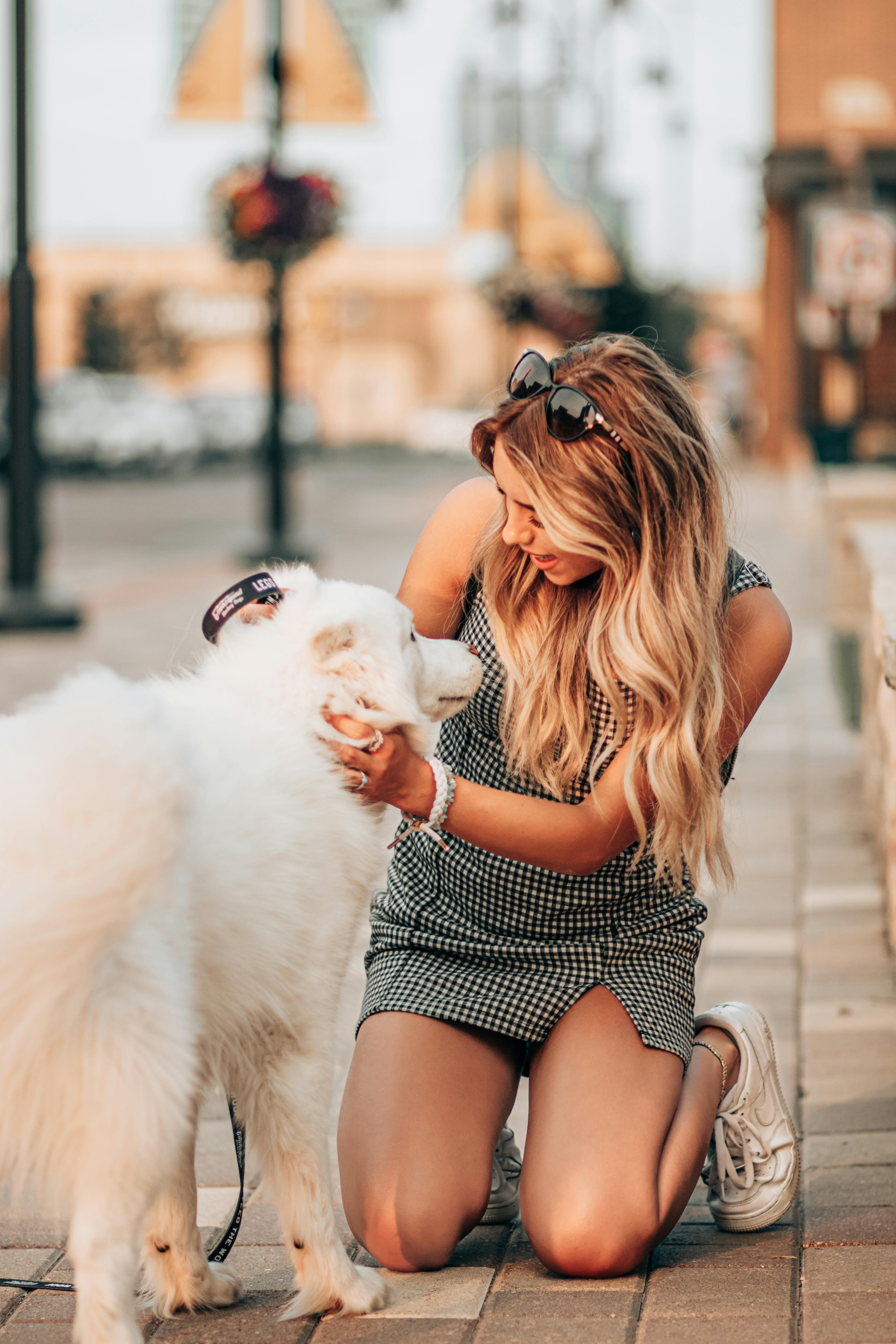 Caucasian woman kneeling on a paved street, playfully interacting with her fluffy white dog.