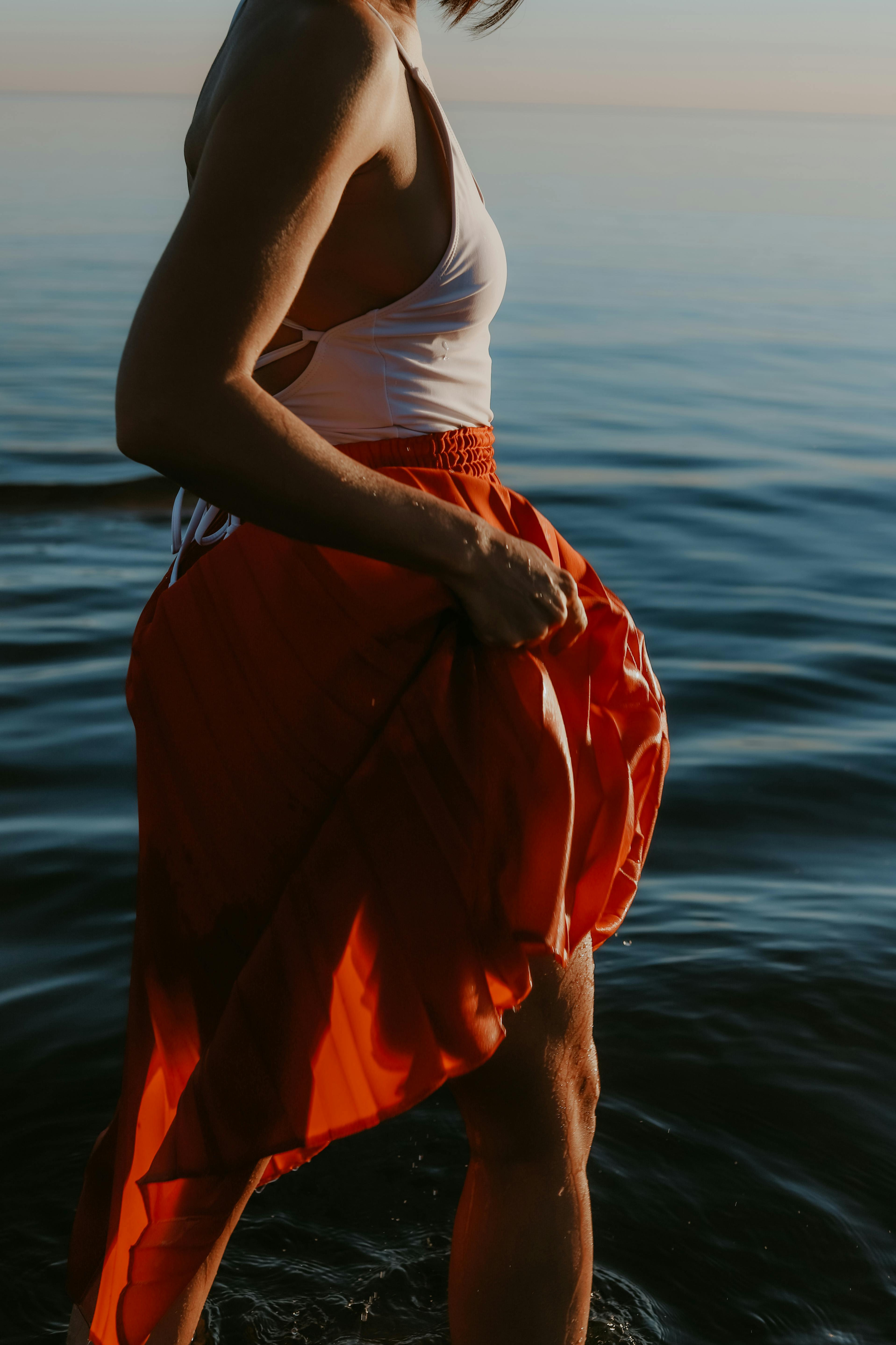 A woman standing in calm ocean waters, wearing a white top and red skirt, at dusk.