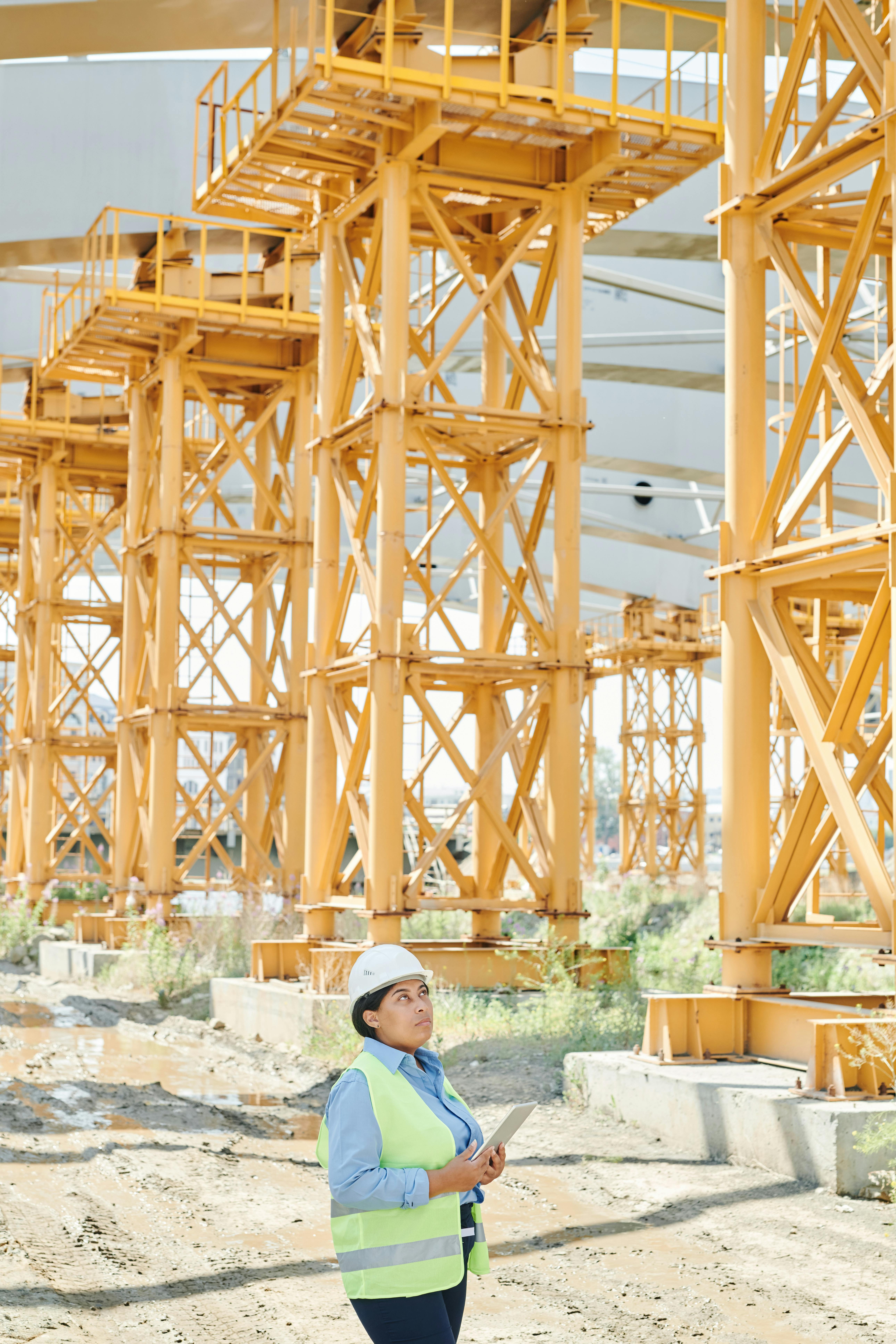 woman in green vest and white hardhat standing near yellow steel structures