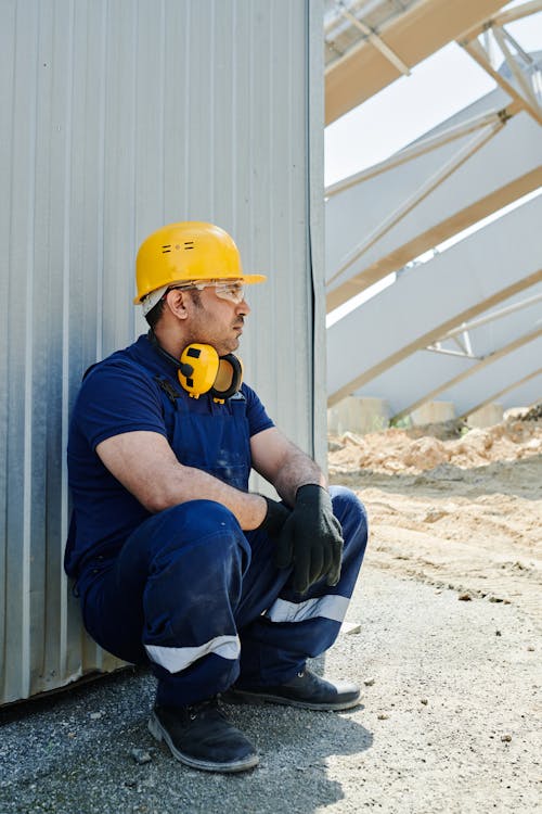 Man in Bue Workwear and Yellow Hardhat Sitting Near a Metal Wall