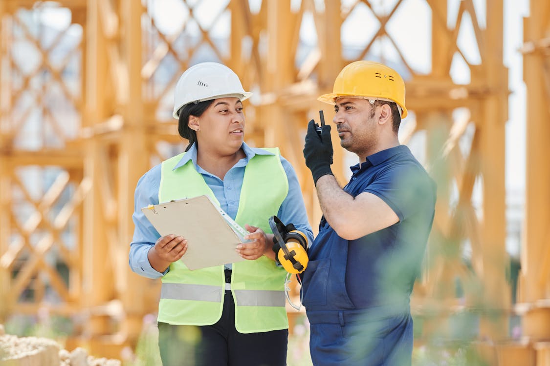 Free Engineers in safety gear discussing a construction project outdoors with visible scaffolding. Stock Photo