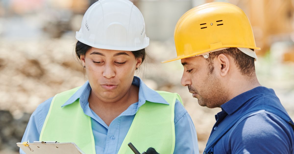Two construction workers in hard hats and reflective vests discuss site safety plans.