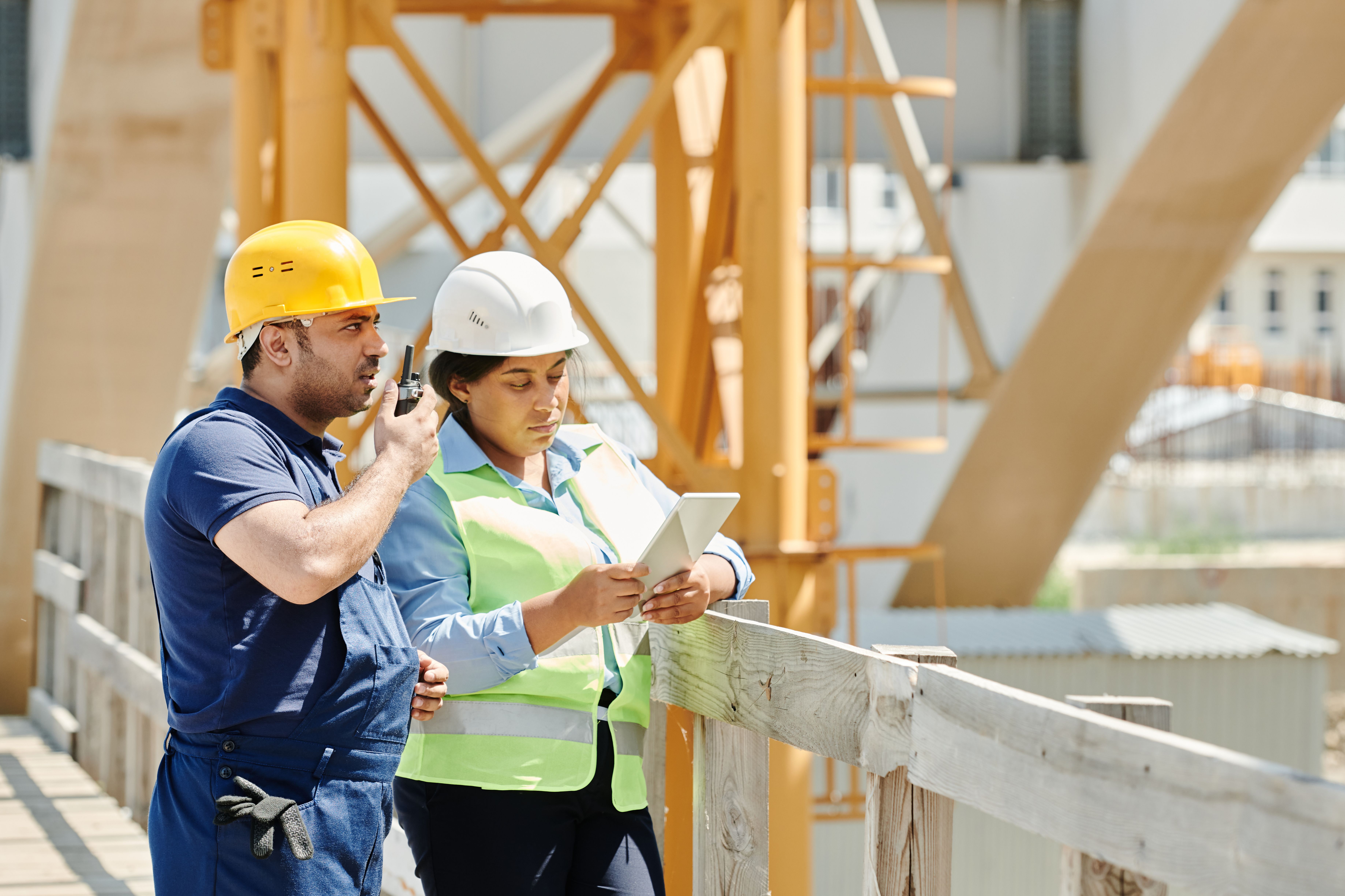 Two construction workers in hard hats discuss plans using a tablet and walkie talkie on-site