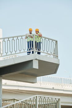 Two construction workers in hard hats inspecting plans on an elevated structure.