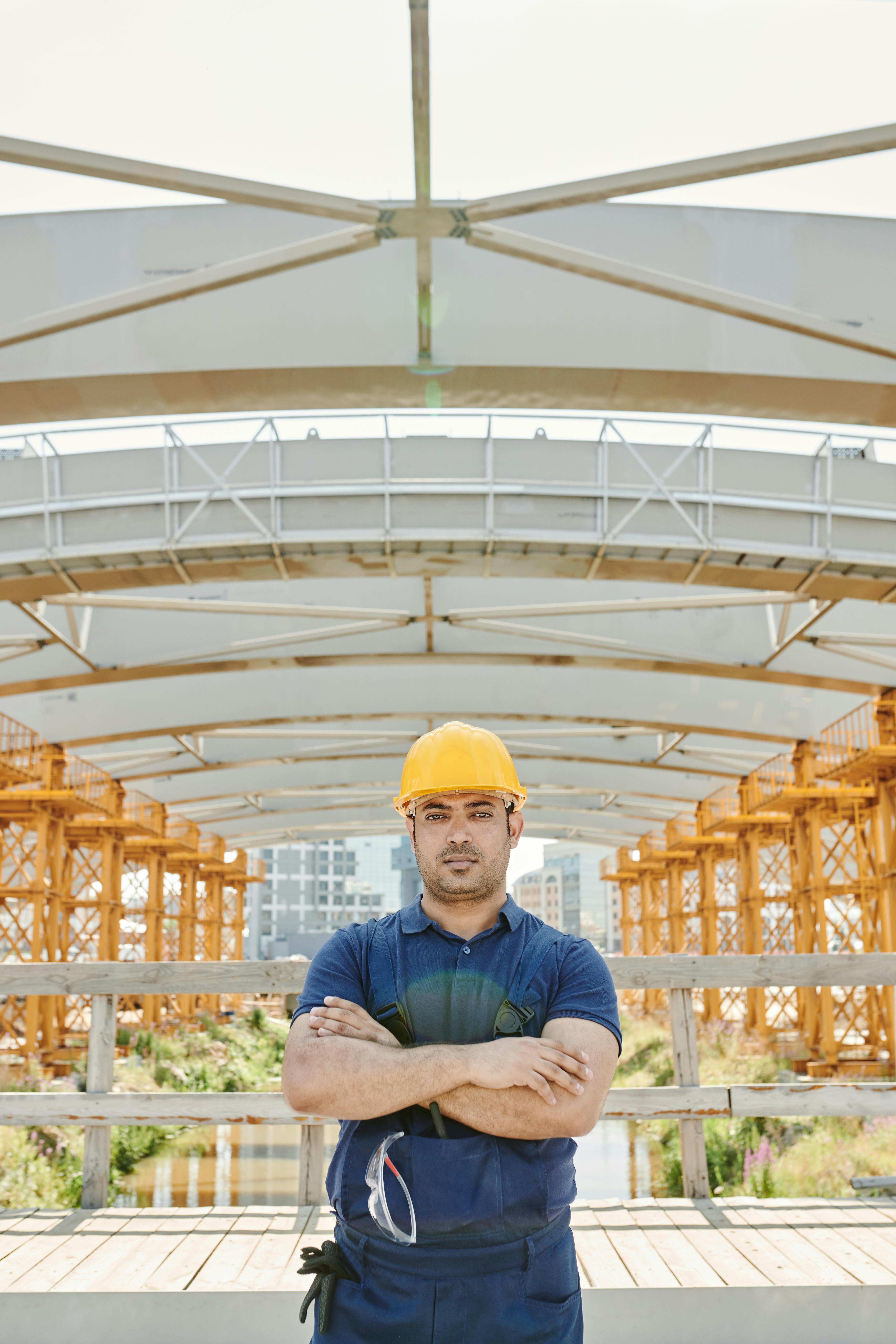 a man with yellow hardhat standing under a truss with crossed arms