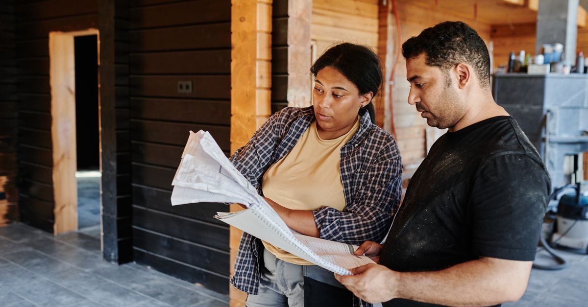 Two construction workers review blueprints inside a wooden structure, discussing plans.