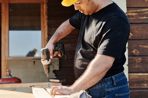 Free A construction worker wearing a helmet uses a power drill on a wooden plank at an outdoor construction site. Stock Photo