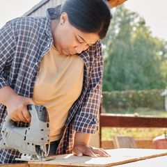 Woman in a checked shirt cutting a board with a grey jigsaw at an outdoor workbench on a sunny day