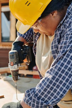 Woman in a hard hat using a power drill on wood indoors, showcasing focused craftsmanship and skill.