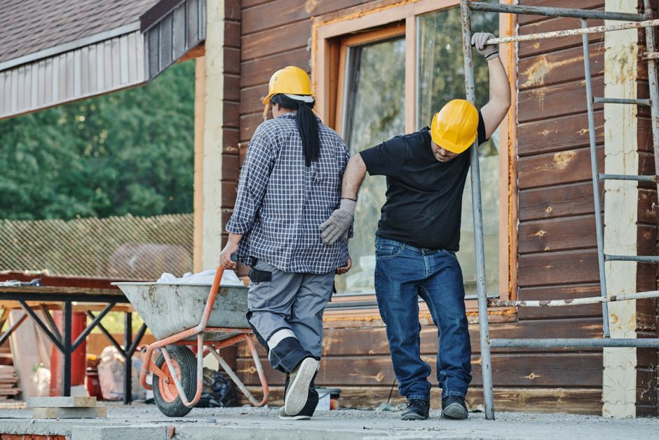 Dos trabajadores de la construcción con cascos trabajan en un sitio al aire libre, uno con una carretilla.