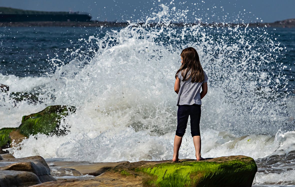 A Girl in Stripes Shirt Standing on the Rock Formation in the Shore