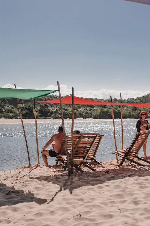 Free People enjoying a sunny day under colorful sunshades on a sandy beach by the water's edge. Stock Photo