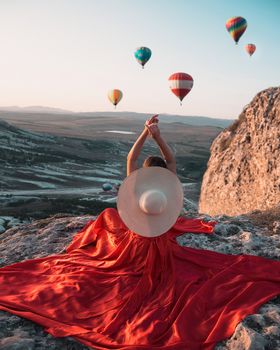 A woman in a red dress and hat enjoys a scenic view with hot air balloons over a rocky landscape.