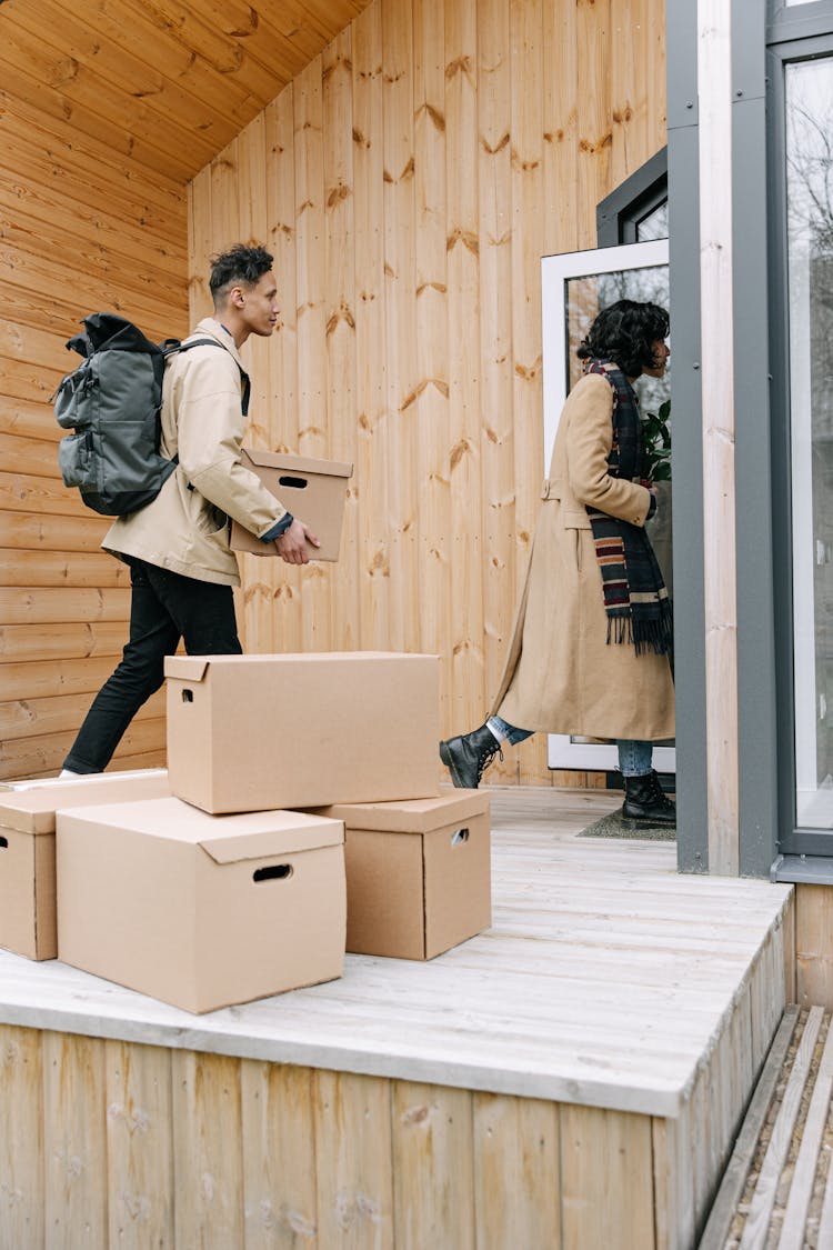 A Man And Woman Carrying Cardboard Box While Going Inside The House