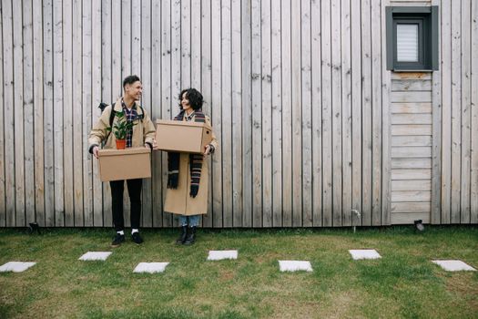 Happy couple carrying boxes and a plant, moving into a new home against a wooden wall.